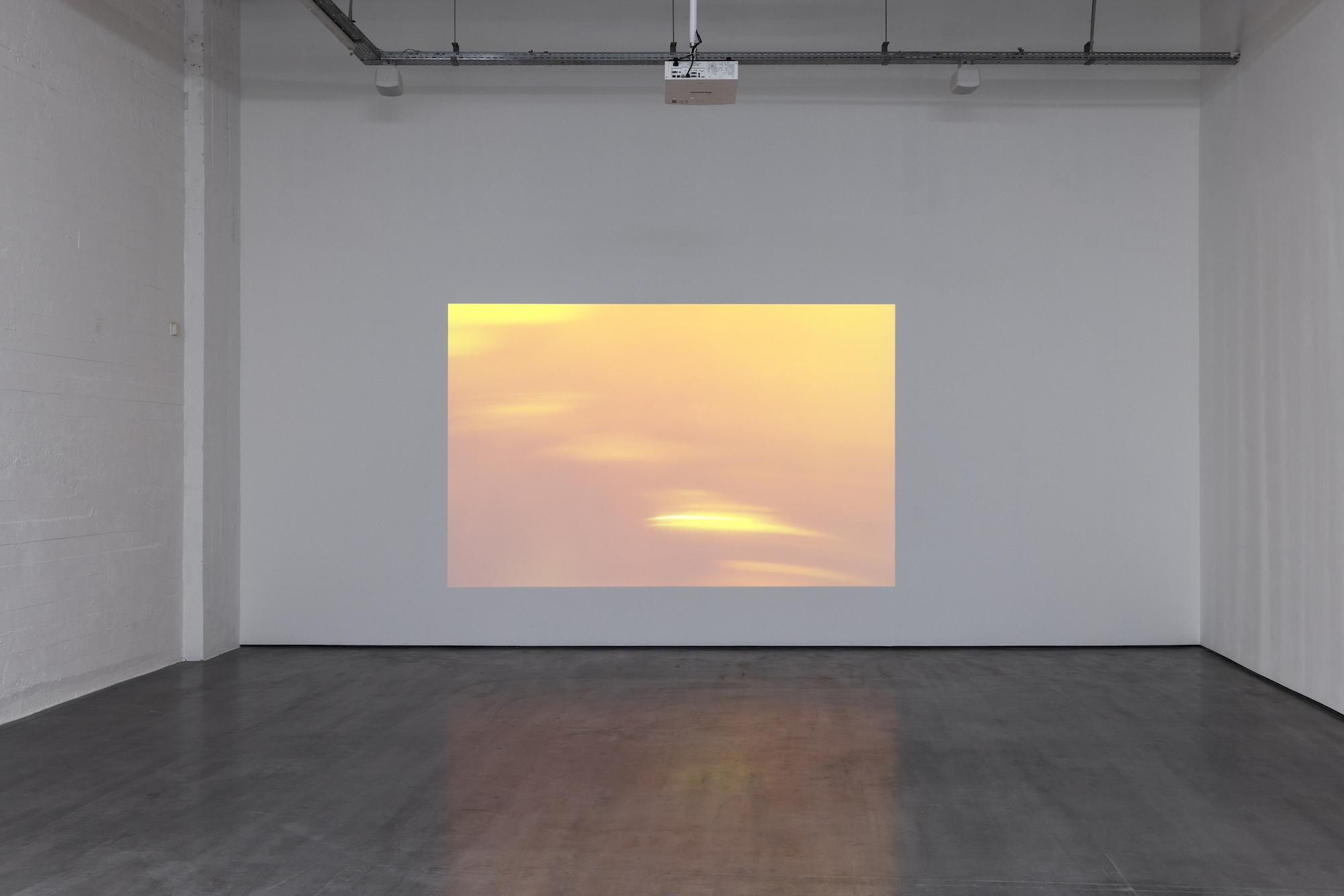 A large rectangular projection of blurred yellow, orange, and pink light fills a white gallery wall and casts a soft reflection across the concrete floor.