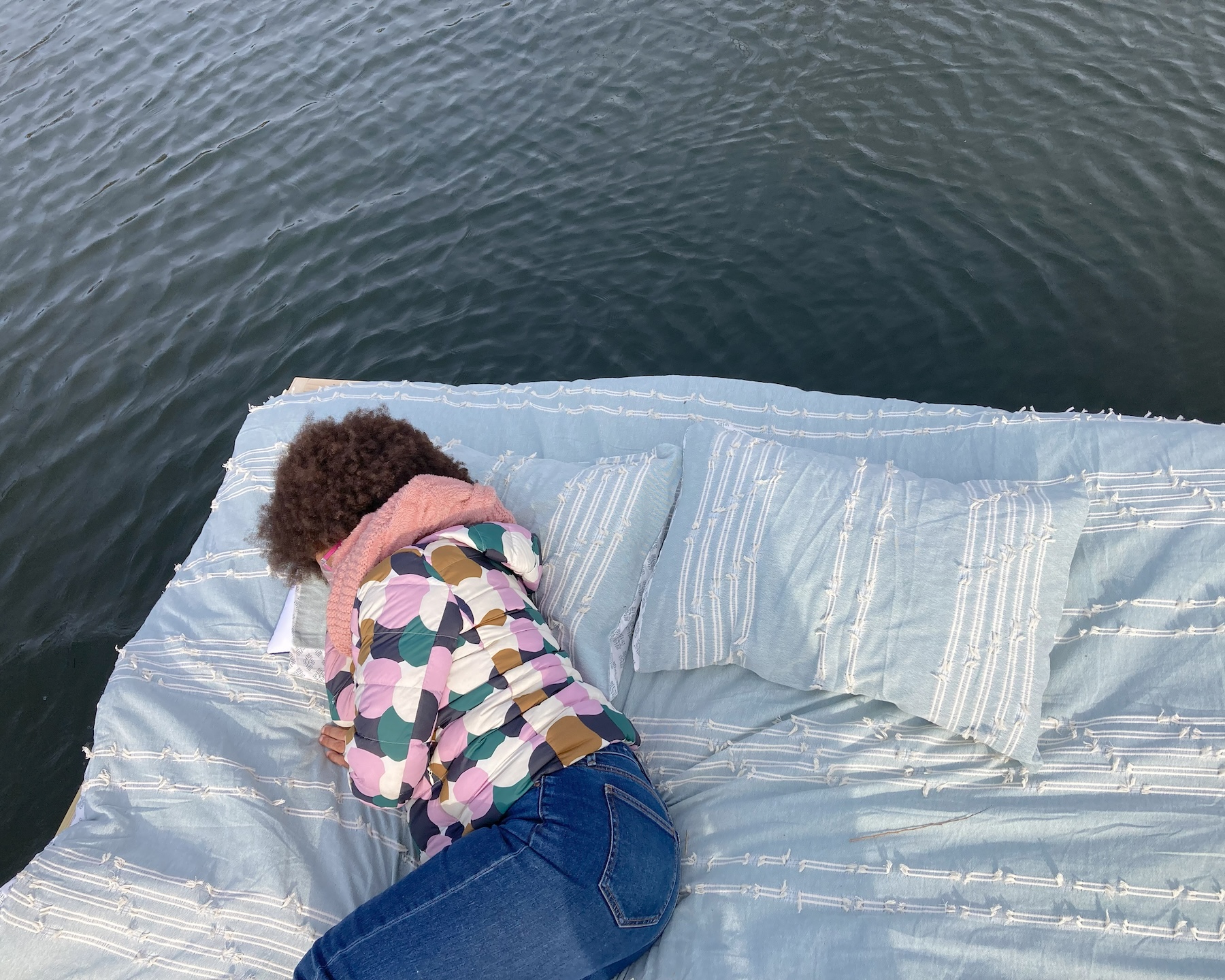Person curled on a pale blue cushioned platform floating on dark water.