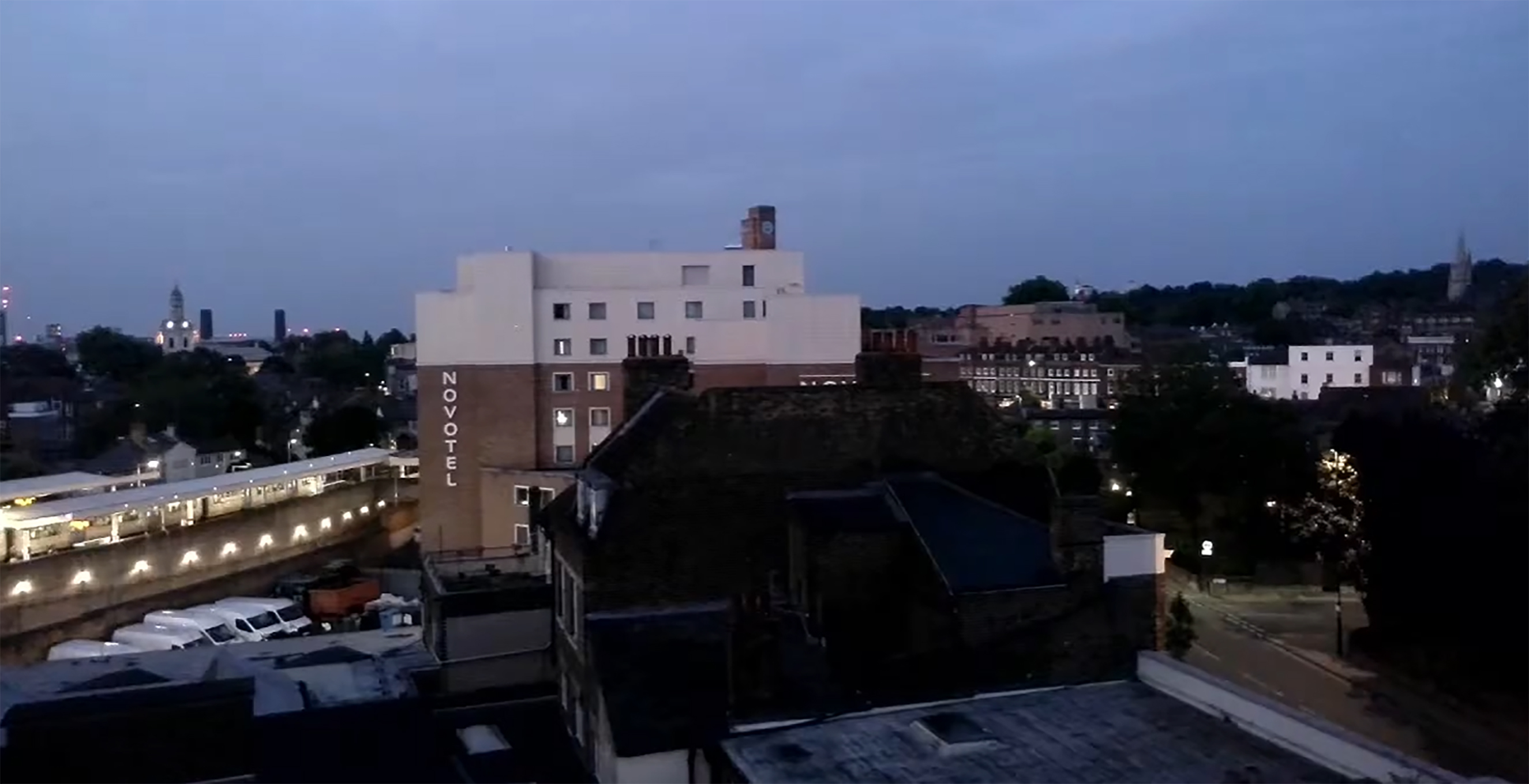 Dusk view over an urban neighborhood, with rooftops and buildings silhouetted beneath a blue-gray sky and a Novotel building visible in the distance.