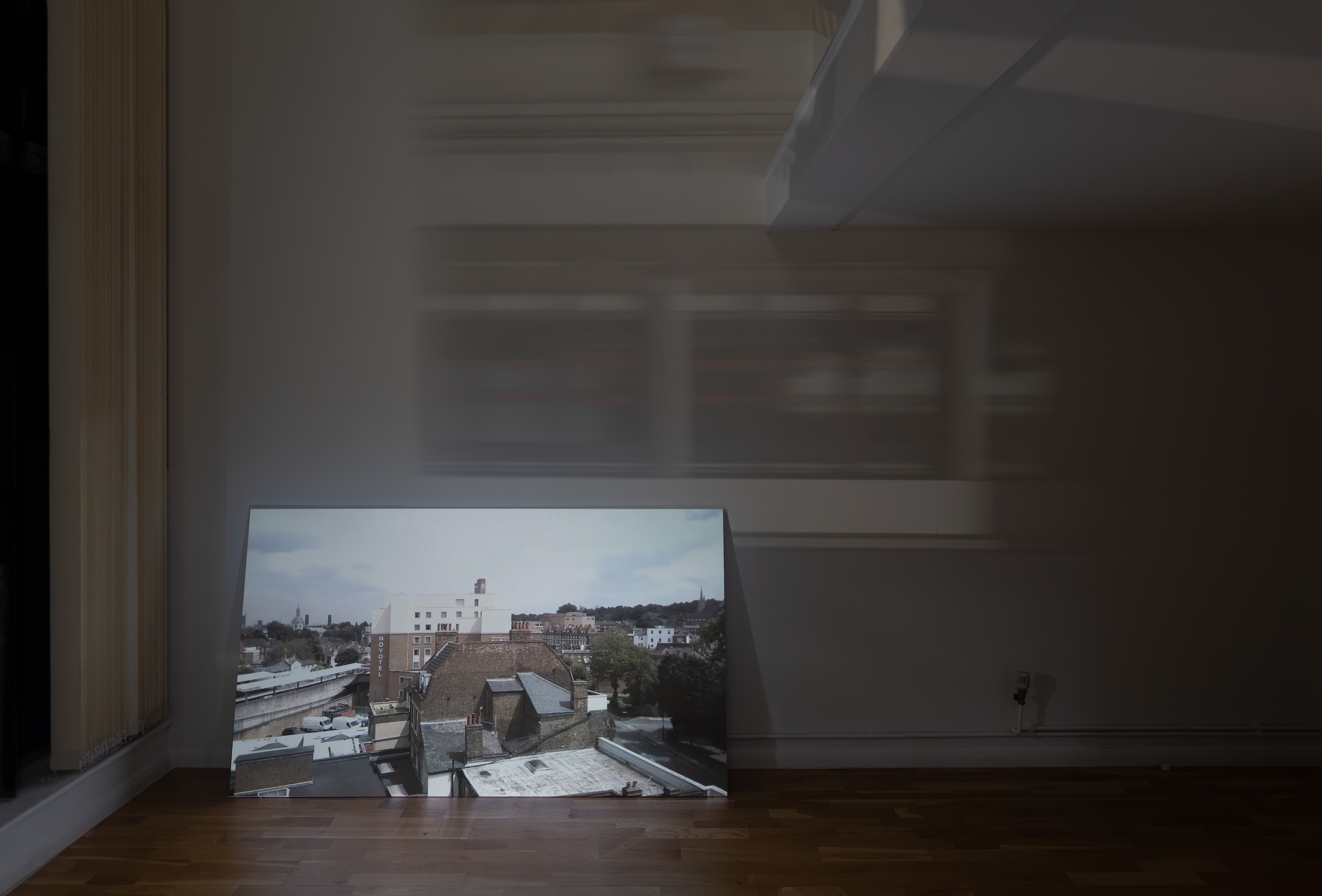 Installation view of a darkened room, where a video projection showing the surrounding cityscape leans against the wall. Blurred bands of light and shadow stretch across the wall above it.