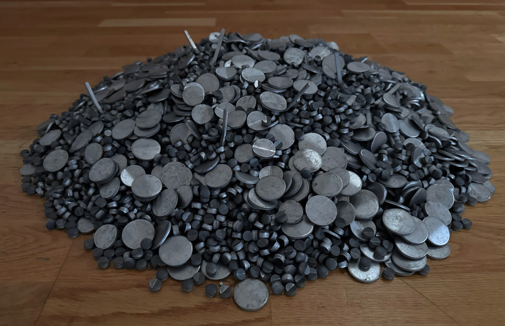Close-up of a large mound of dark gray metal discs, cylinders, rods, and irregular pieces piled on a wooden floor.