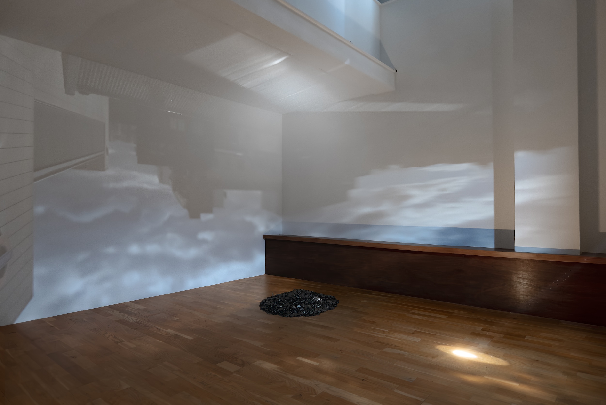 Installation view of a sparsely furnished interior, with hazy projections of architectural forms and clouds cast across white walls. A mound of dark metal pieces rests on the wooden floor at center.