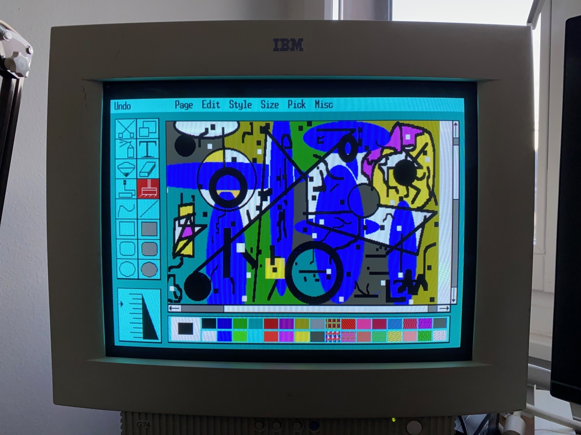 Close-up of a vintage IBM CRT monitor displaying a colorful abstract digital composition within an early graphics-editing program. The screen features overlapping geometric shapes, bold black lines, and patches of blue, green, yellow, and purple, surrounded by a turquoise interface with drawing tools and color palettes.