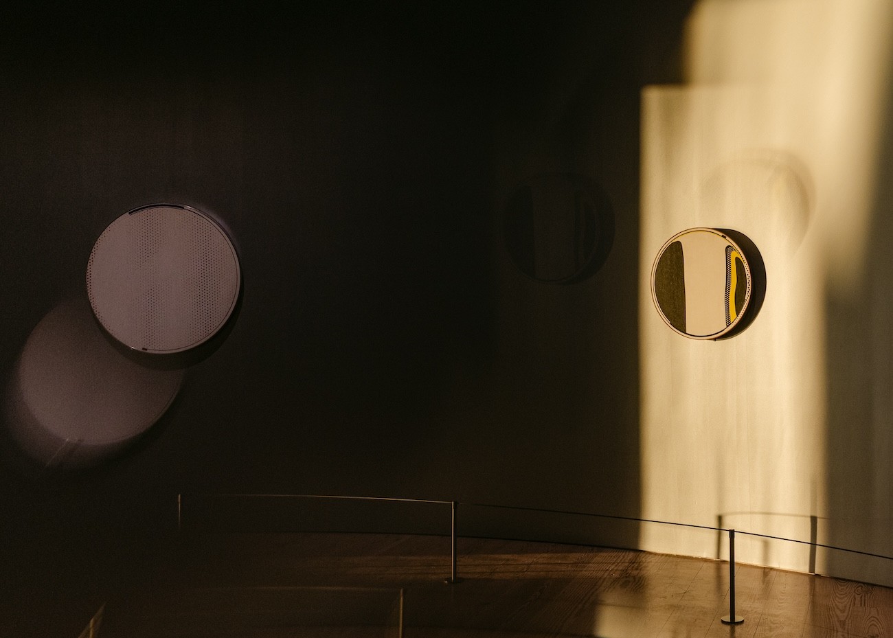 Sunlight cuts across a curved gallery wall displaying Lichtenstein’s circular mirror paintings, leaving the dotted painting at left in shadow and illuminating a yellow-and-black work at right.