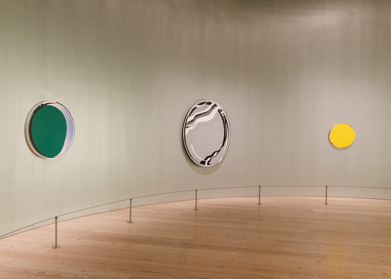 Three of Lichtenstein’s mirror paintings hang above a wooden floor on a curved gray wall: a green circle, a black-and-white oval, and a small yellow circle.