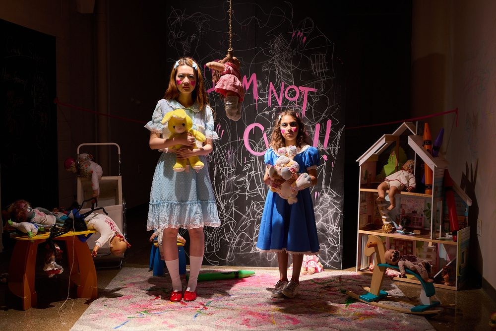 Two performers hold small figures amid a densely furnished theater set with a dollhouse, hanging objects, and a chalkboard.