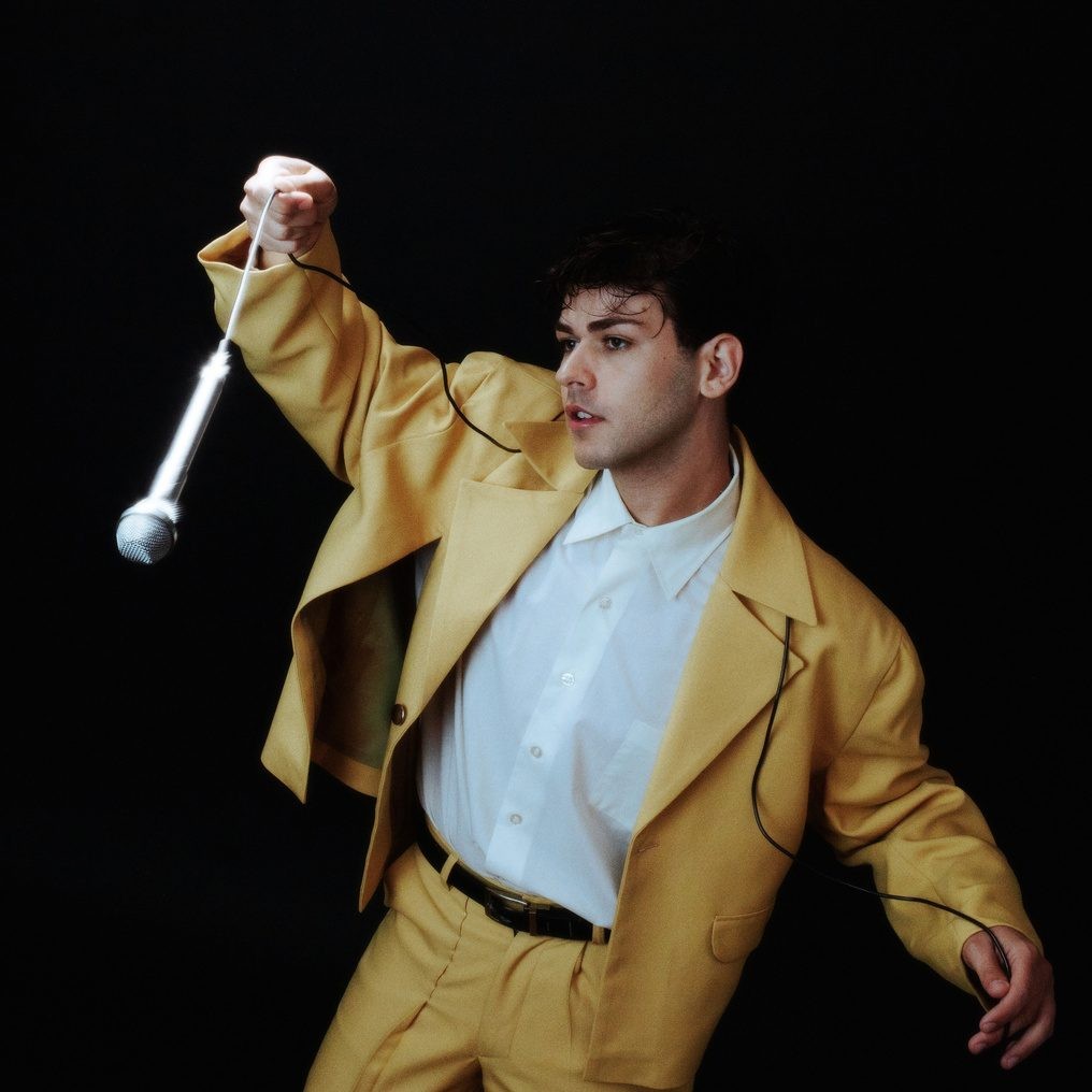 A performer in a mustard-yellow suit holds a microphone by its cord against a black background.
