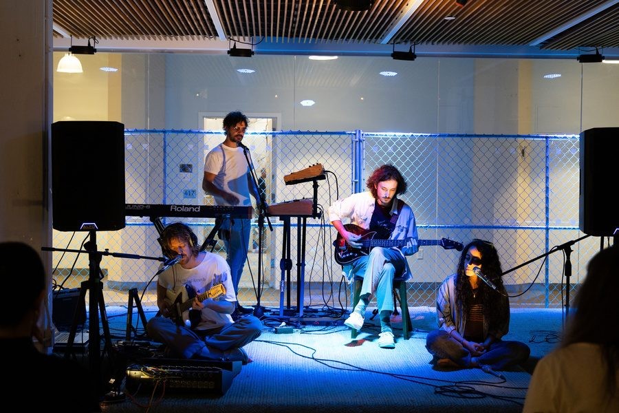A small ensemble performs on a blue-lit stage with a keyboard, guitar, and seated performers in front of a chain-link partition.