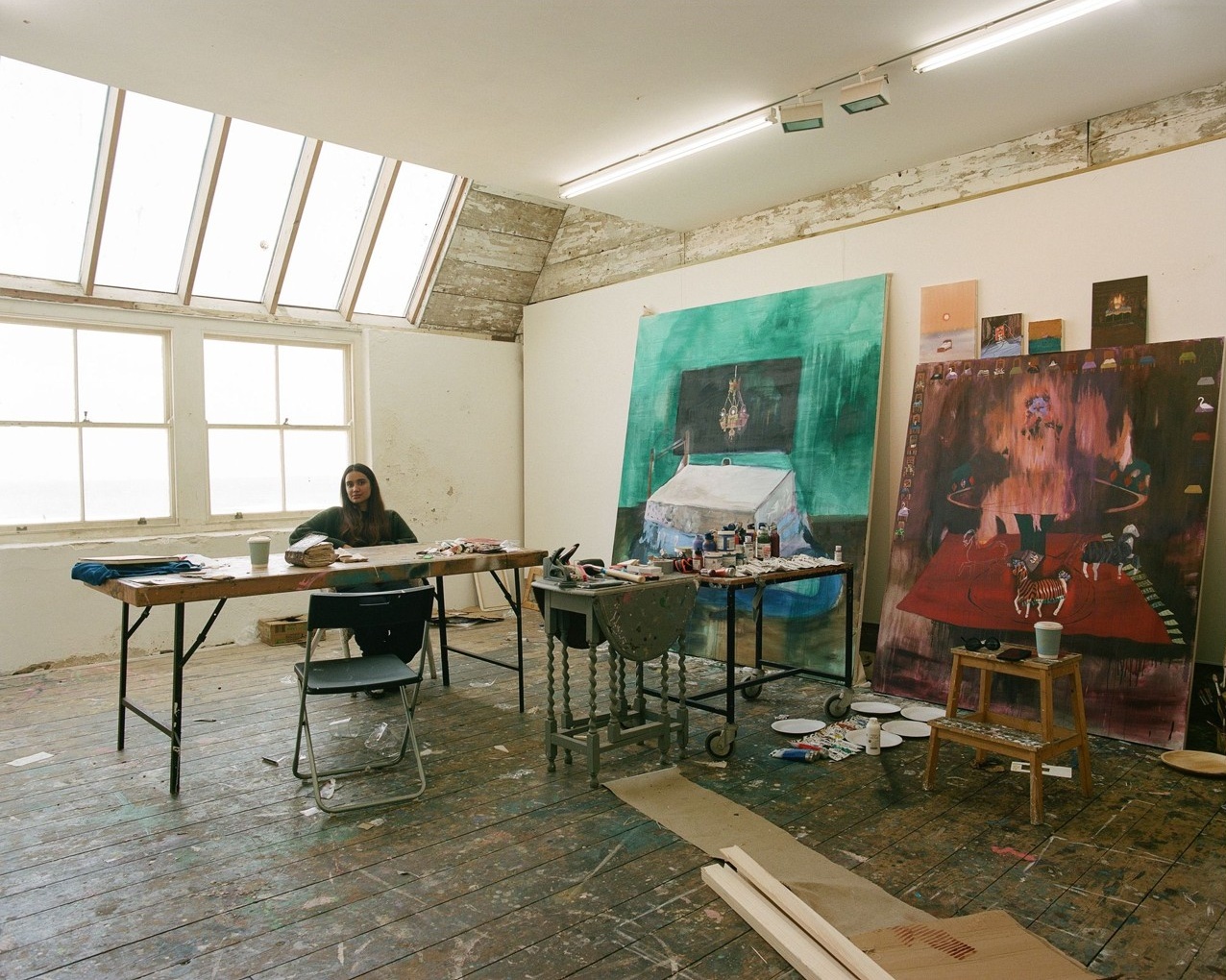 Noorain Inam works at a table in a bright Porthmeor studio, with large windows, skylights, and colorful paintings leaning against the walls.