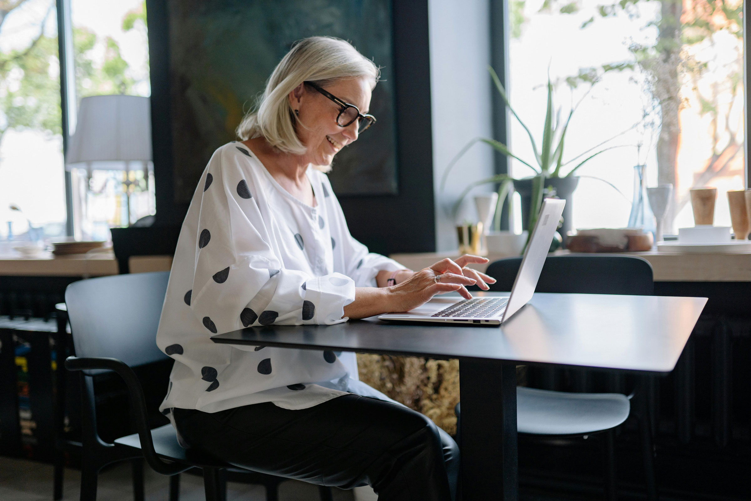 Smiling elderly woman with glasses typing on a laptop at a black table in a well-lit room with plants and decorative items.
