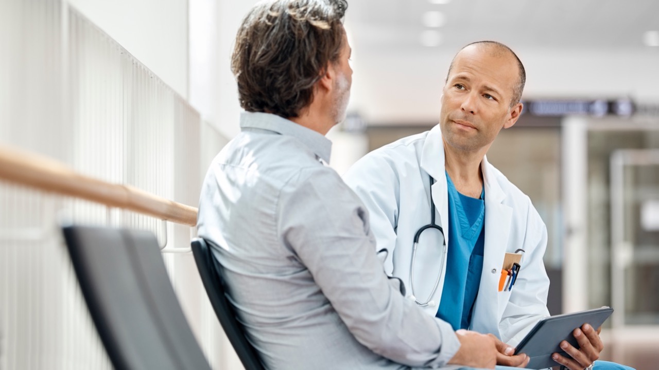 Doctor in white coat with stethoscope consulting a seated patient in a medical facility.