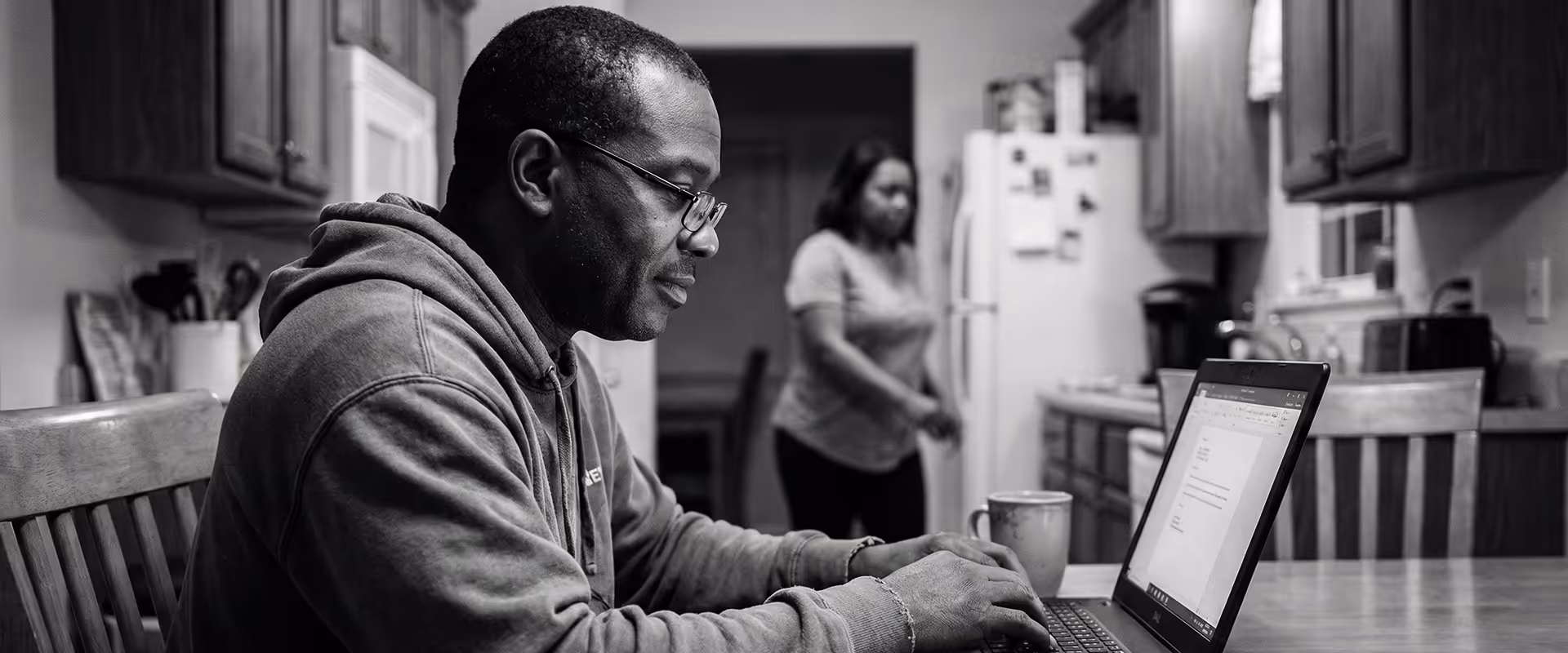 Man in glasses and hoodie typing on a laptop at a kitchen table with papers and a coffee mug.