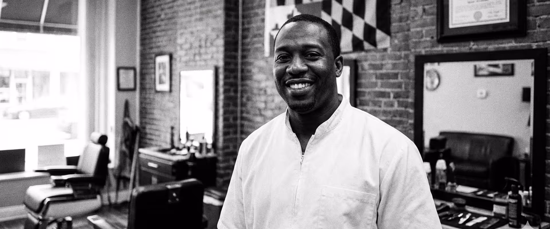 Smiling barber in white coat standing inside a barbershop with chairs, brick walls, and Maryland flag.