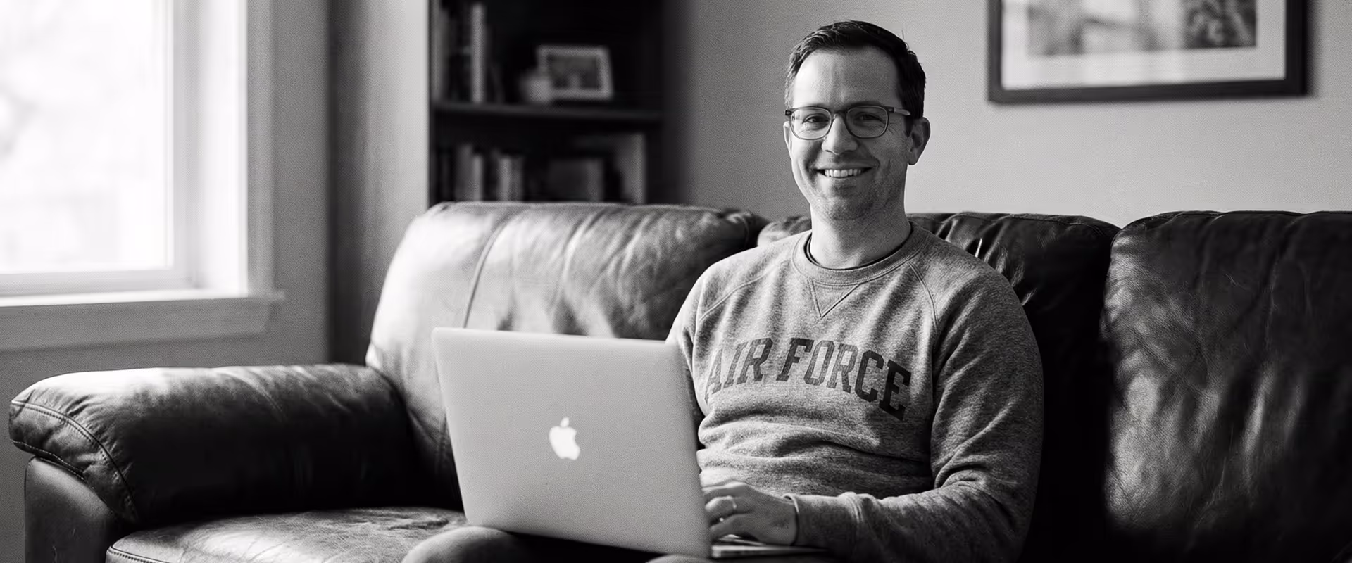Smiling man wearing glasses and an Air Force sweatshirt sitting on a leather couch with a laptop.