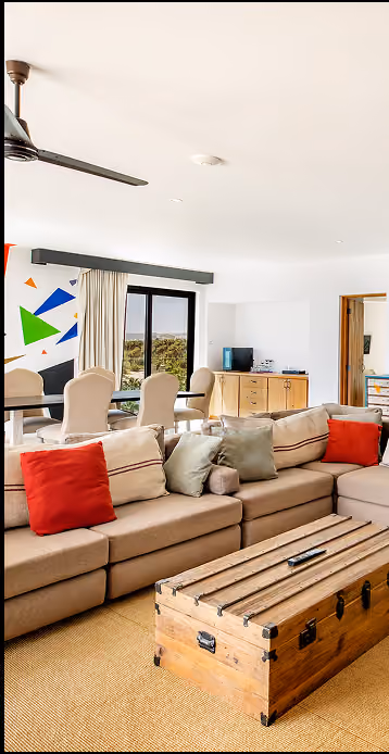 Luminoso salón con sofá modular beige realzado por cojines rojos y verdes, mesa de centro de madera, comedor con sillas tapizadas y gran ventana con vistas.