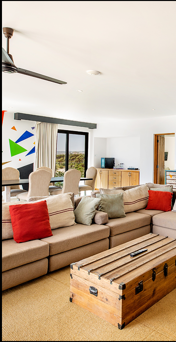 Bright living room with beige sectional sofa accented by red and green cushions, wooden chest coffee table, dining area with upholstered chairs, and large window with a view.