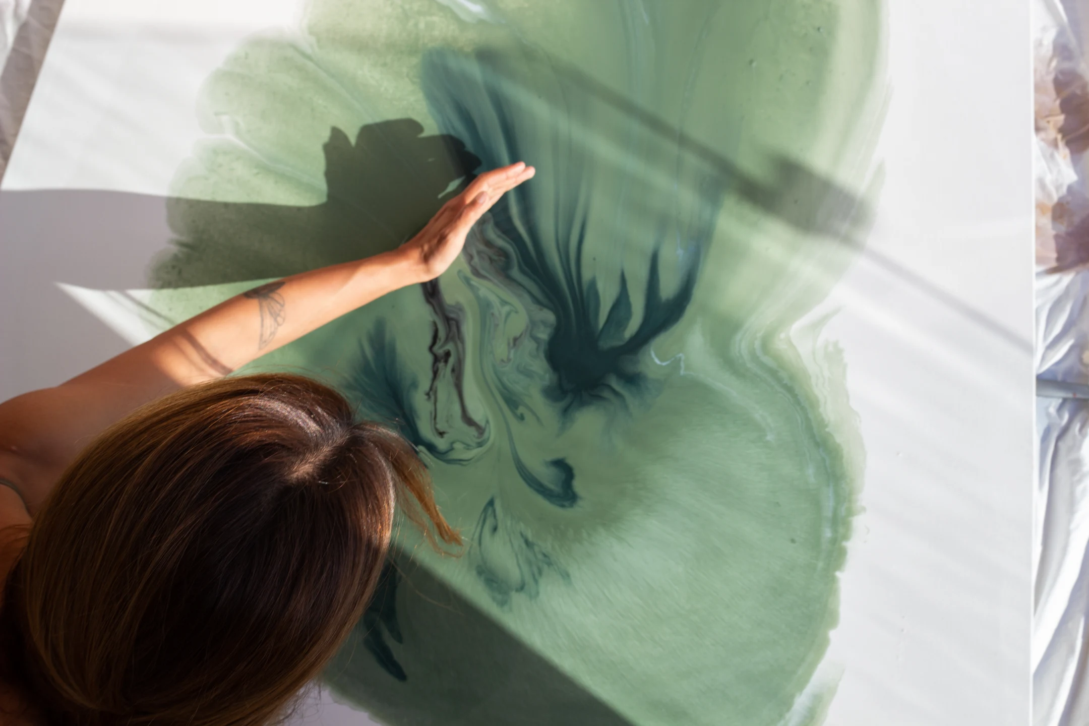 Person with tattooed arm spreading green and dark blue fluid paint on a white canvas.