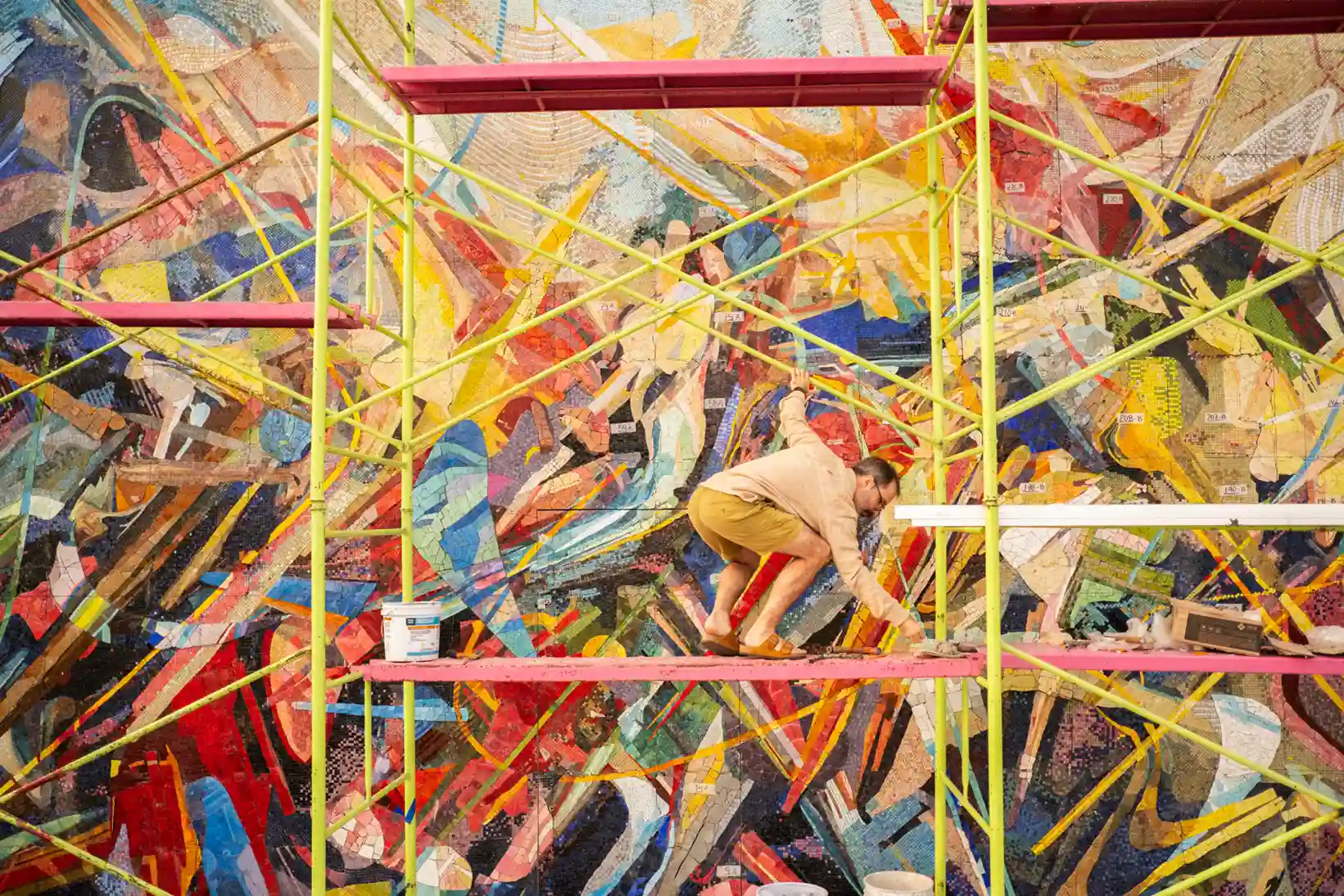 Man crouching on pink scaffolding while working on a vibrant, colorful mosaic mural background.