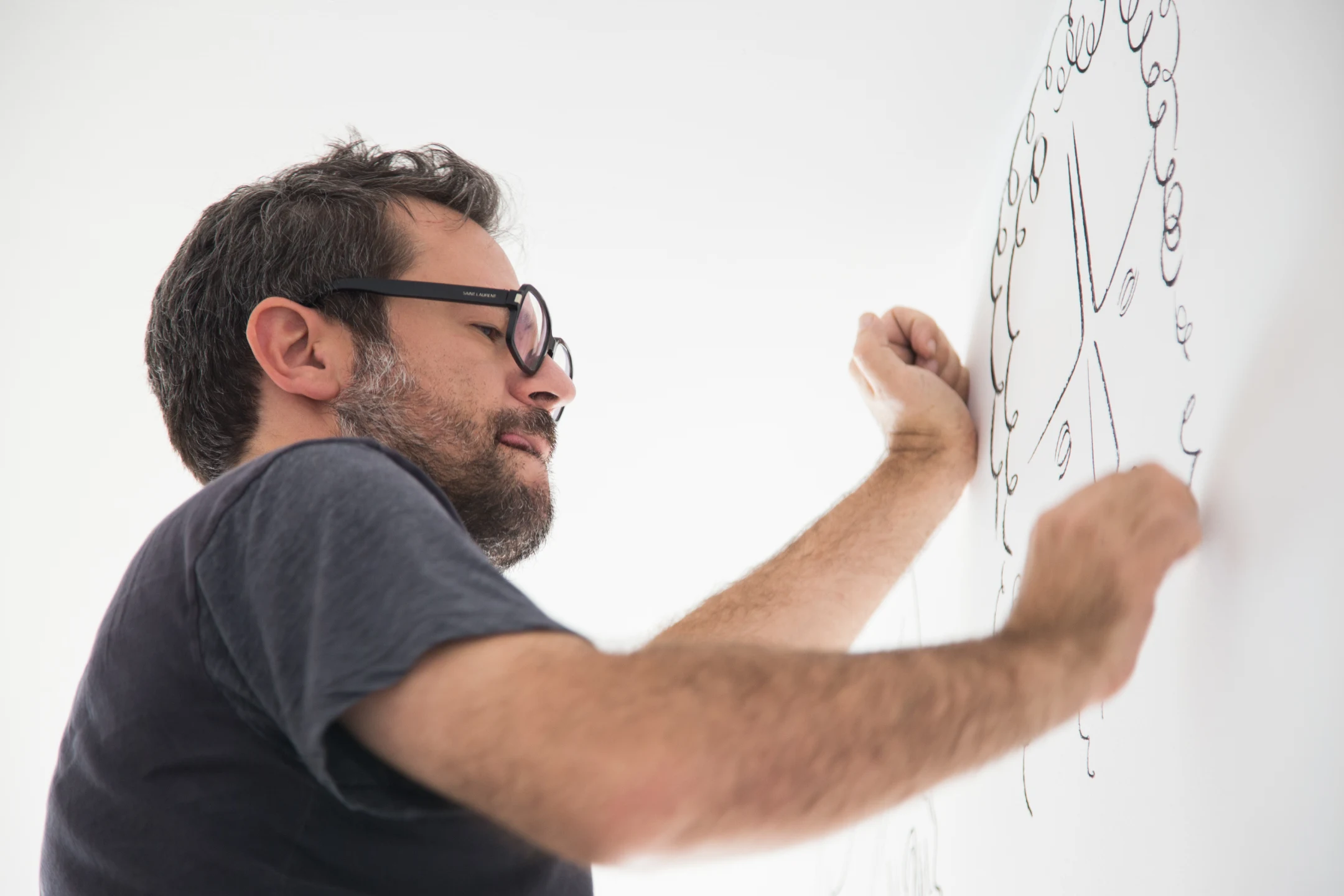 Man wearing glasses drawing a large face with curly hair on a white wall.