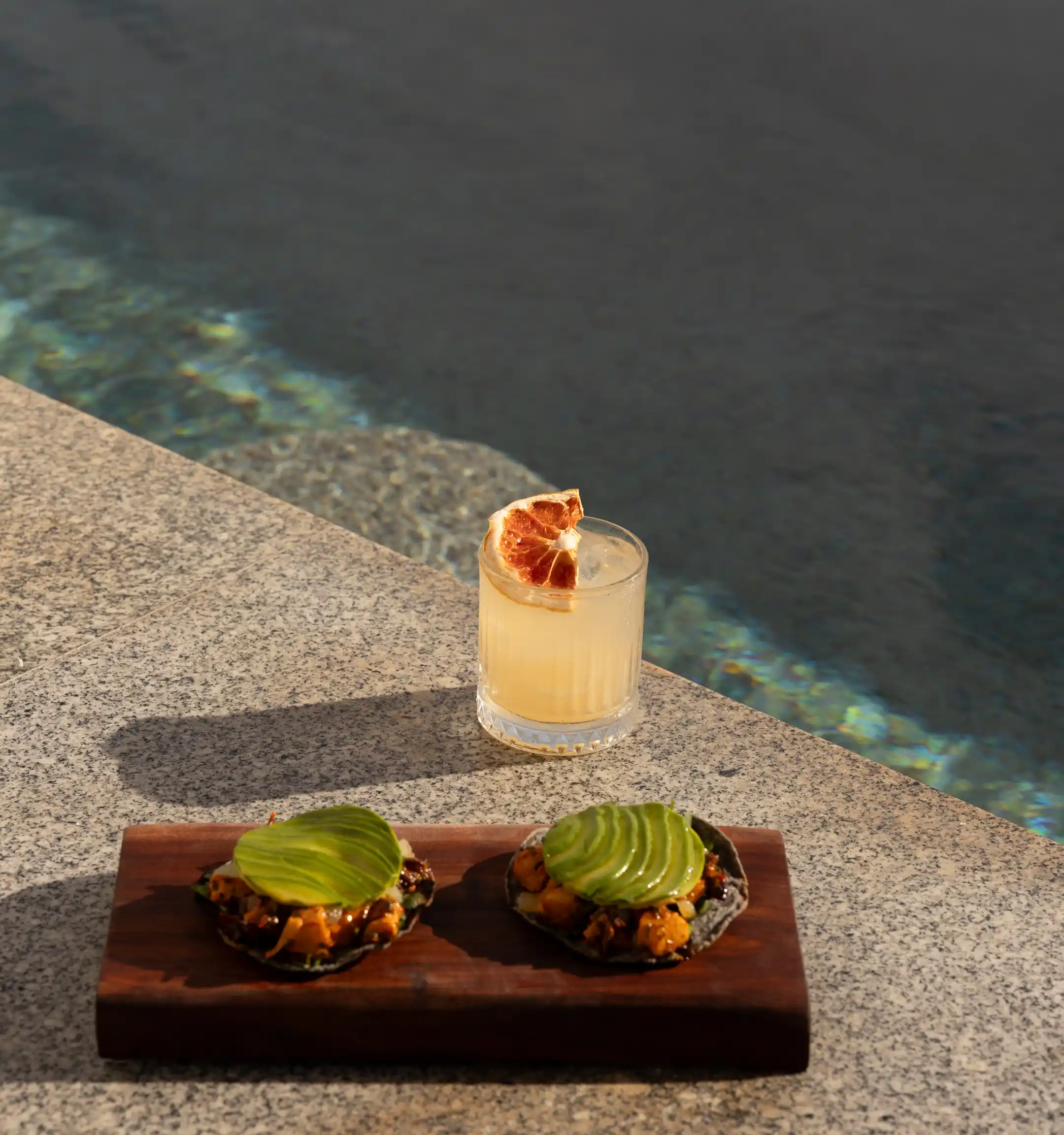 Two appetizers topped with sliced avocado on a wooden tray next to a cocktail glass with a dried citrus garnish near a pool edge.
