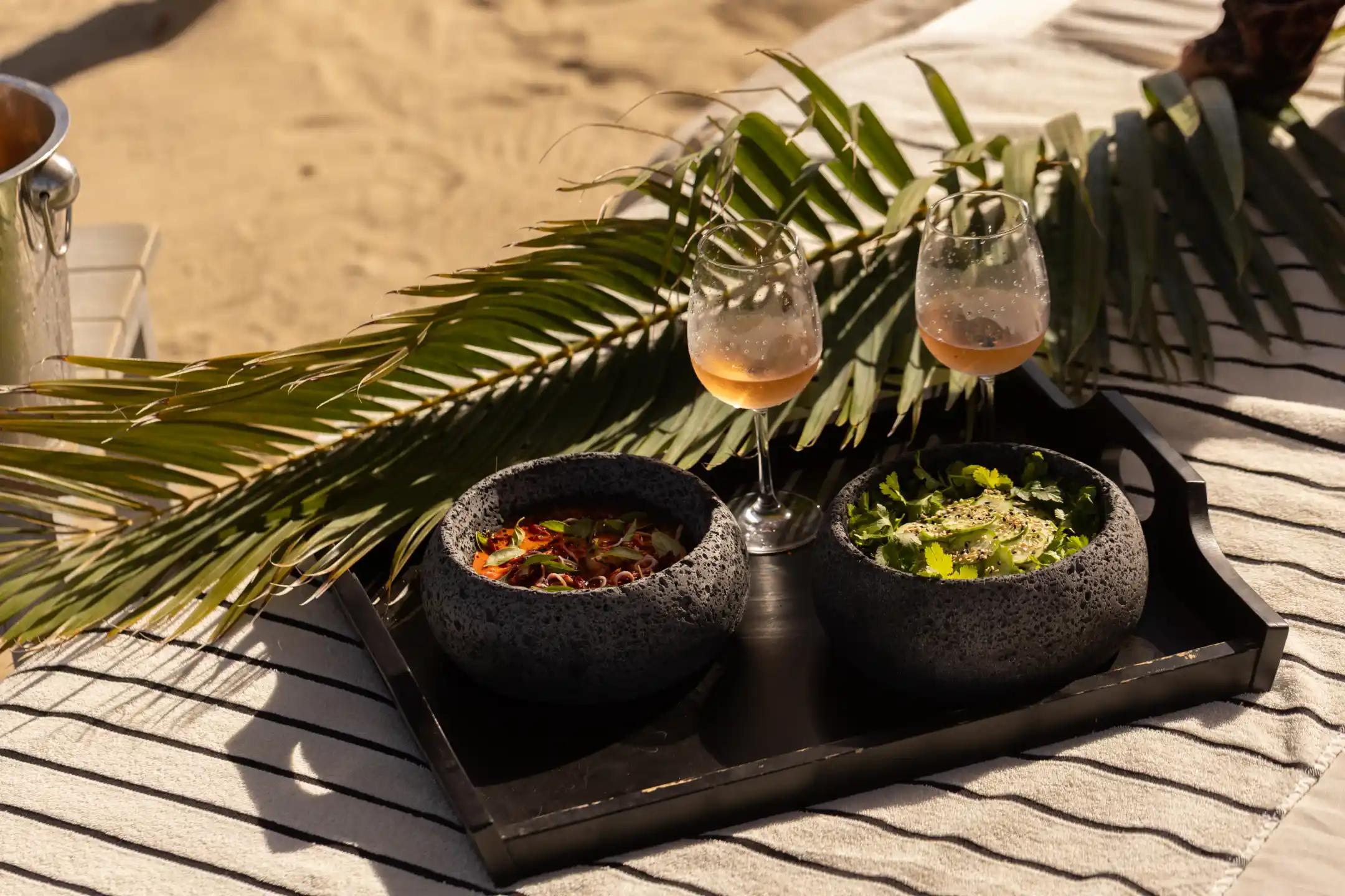 Two glasses of rosé wine and bowls of salad on a black tray with palm leaves, set on a striped cloth on the sand.