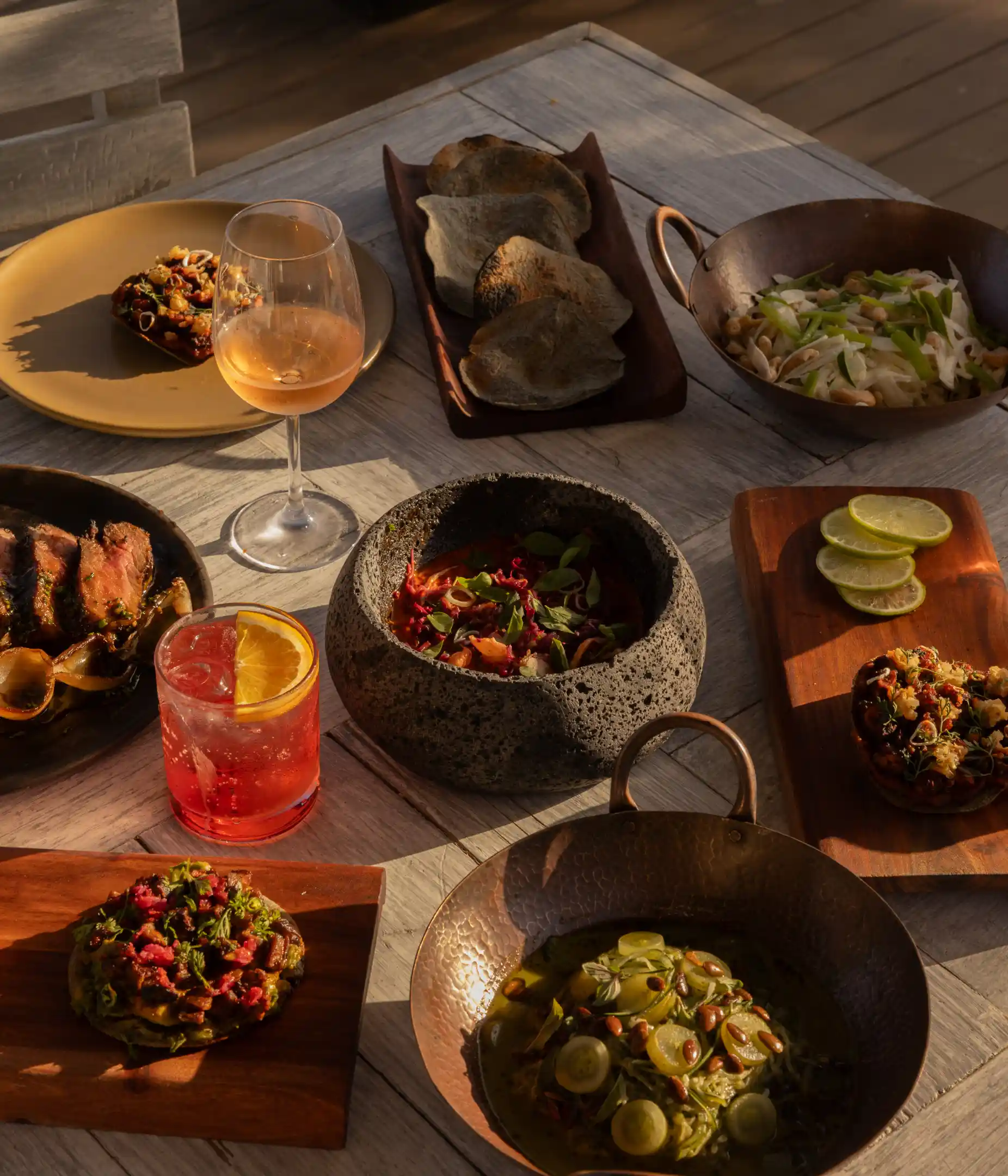 Table with gourmet dishes including steak, salads, appetizers, lime slices, and two drinks in copper and glassware, lit by warm light.