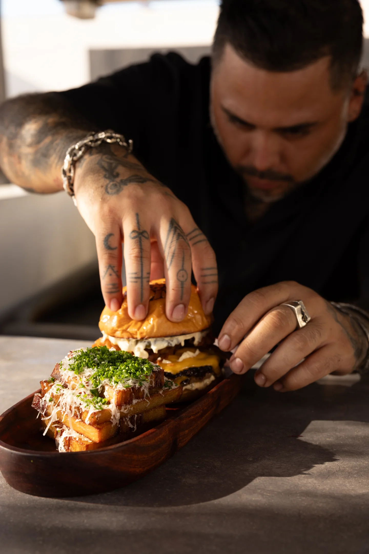 Man with tattoos placing a cheeseburger and loaded fries topped with chives and cheese on a wooden tray.