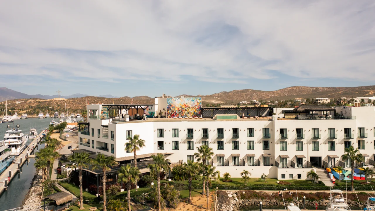 White multi-story hotel building with balconies, rooftop terrace with colorful mural, surrounded by palm trees near a marina with yachts.