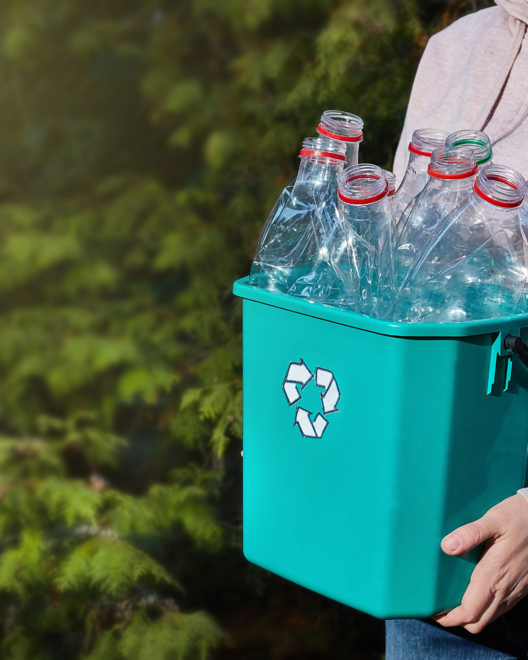 Persona sosteniendo un contenedor de reciclaje de color verde azulado lleno de botellas de plástico vacías al aire libre.