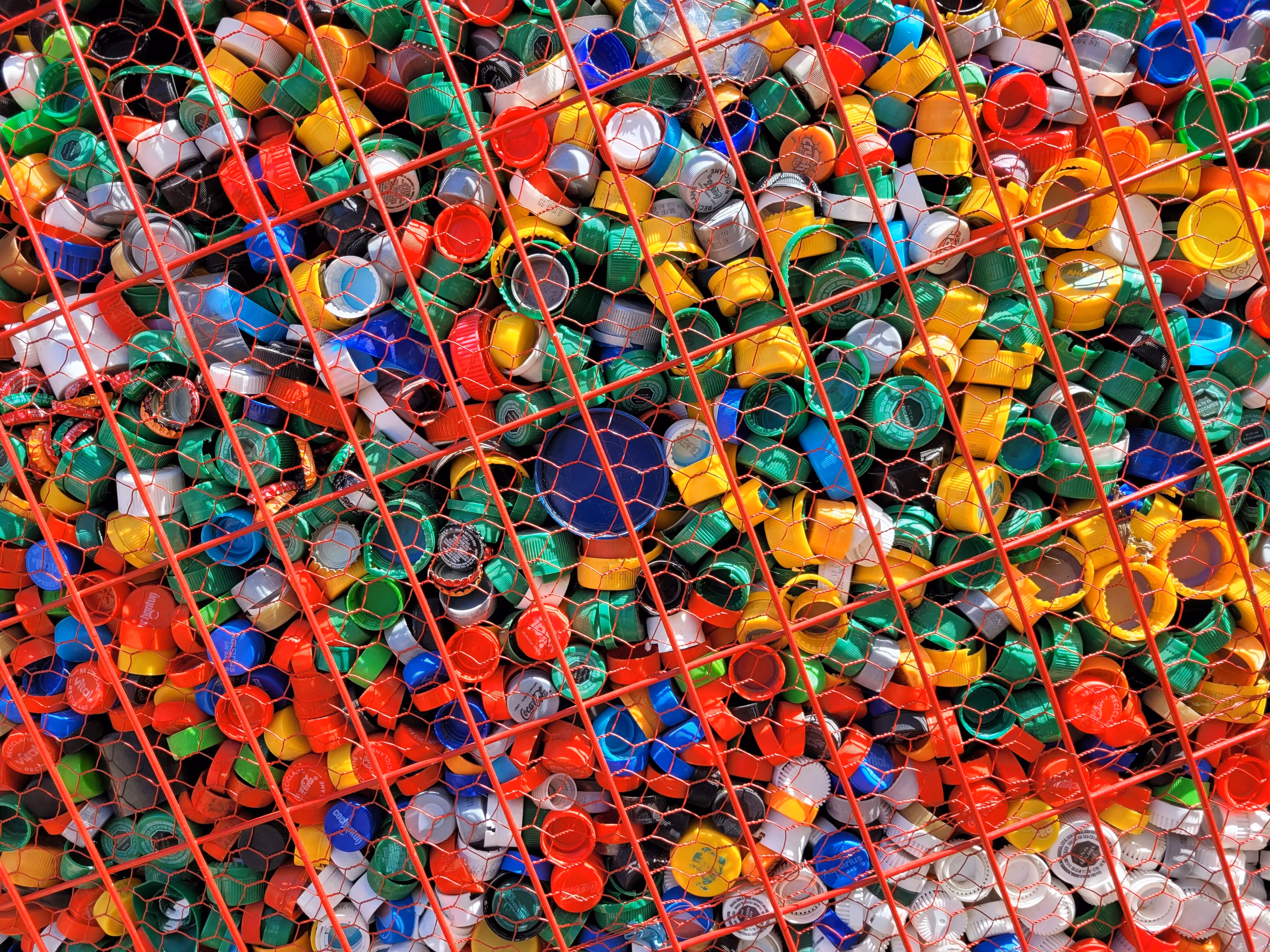 Montón de tapones de botellas de plástico multicolores contenidos detrás de una malla metálica roja.