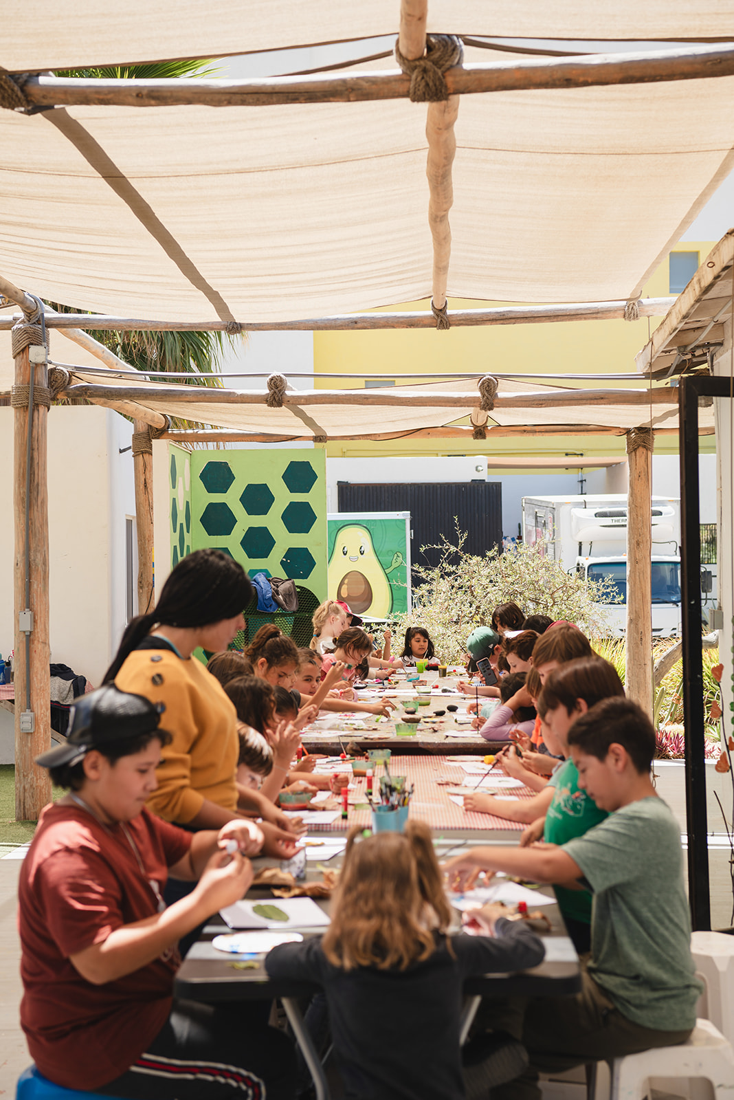 Grupo de niños sentados en una larga mesa al aire libre bajo una lona de sombra, realizando actividades artísticas y manualidades.