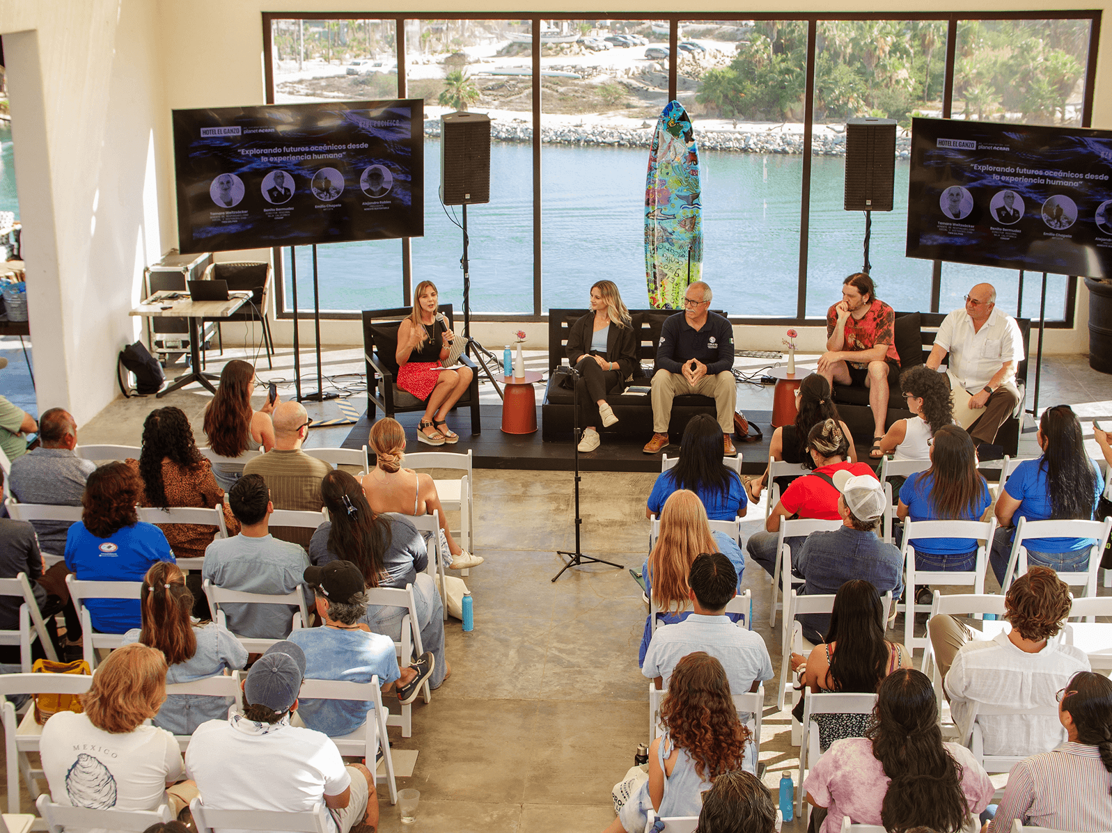 Mesa redonda con cinco ponentes sentados frente a una gran ventana con vistas al agua, con un público que escucha atentamente en una sala luminosa.