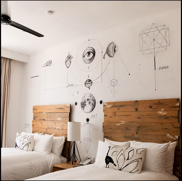Habitación de hotel con dos camas con cabeceros de madera y un mural en la pared con dibujos anatómicos y cósmicos, incluyendo un ojo, un corazón, una oreja, un cráneo de pájaro, las fases lunares y formas geométricas.