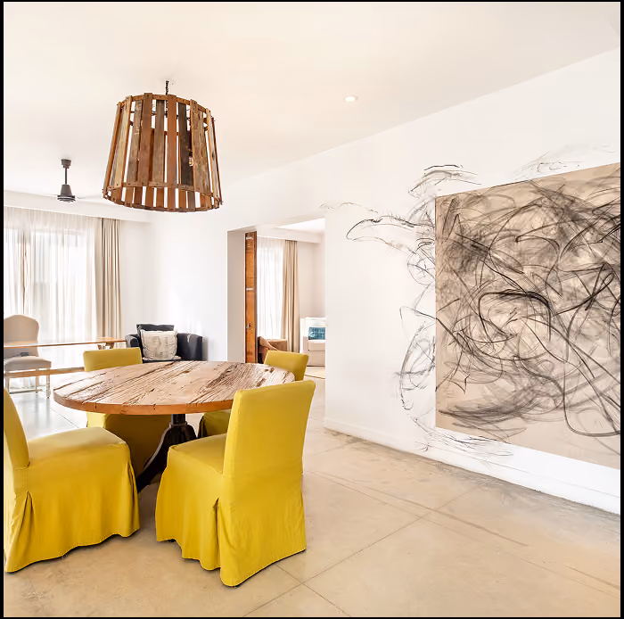Comedor moderno con una mesa redonda de madera rodeada por cuatro sillas tapizadas en amarillo y un gran cuadro abstracto en negro y gris sobre una pared blanca.