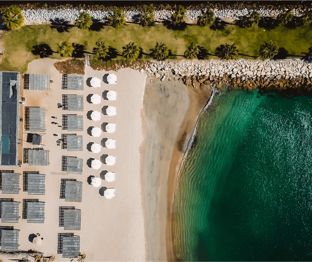 Aerial view of a beach resort with rows of white umbrellas, shaded wooden cabanas, green grass, palm trees, rocky breakwater, and clear turquoise water.