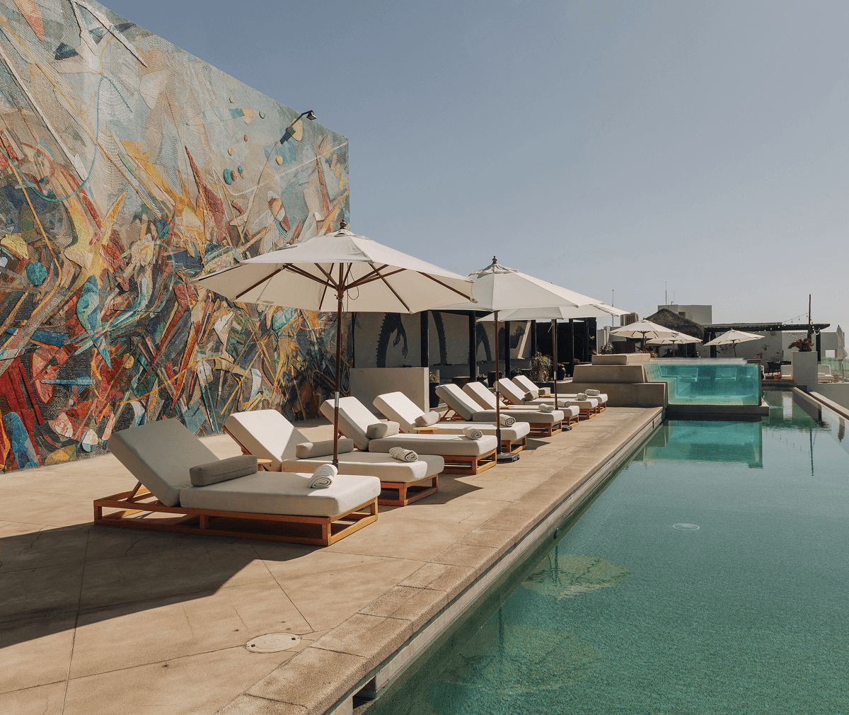 Row of white cushioned lounge chairs with umbrellas beside a rooftop pool and a colorful abstract mural on the wall.