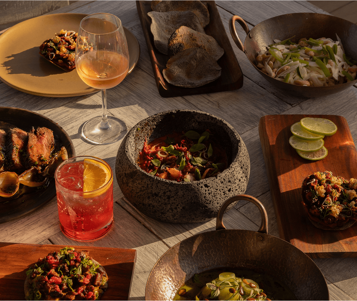 Table with various gourmet dishes including grilled meat, appetizers, lime slices, and two cocktails in glassware under warm lighting.