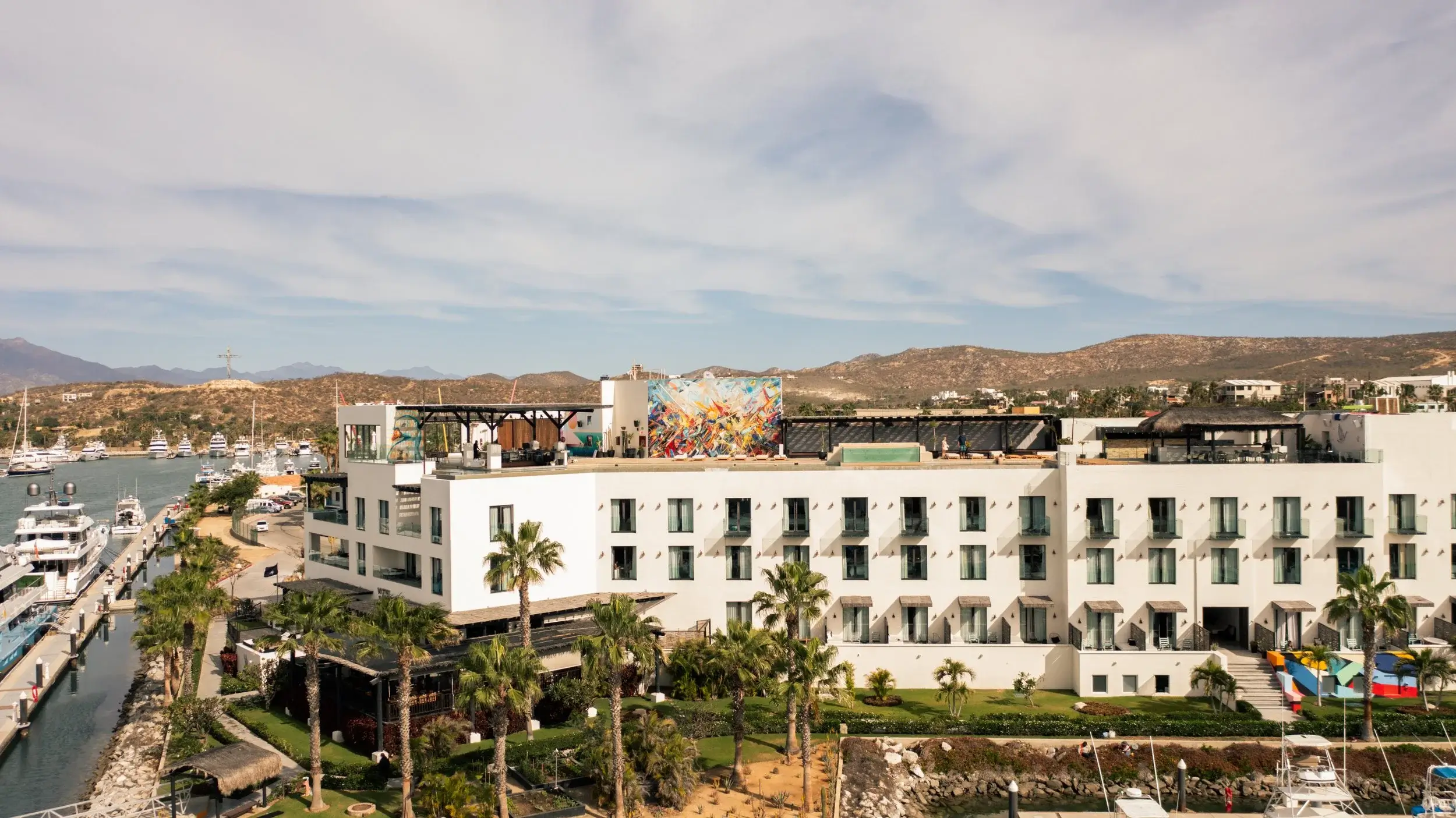 White multi-story hotel building with balconies and rooftop terrace overlooking marina with yachts and arid hills in the background.