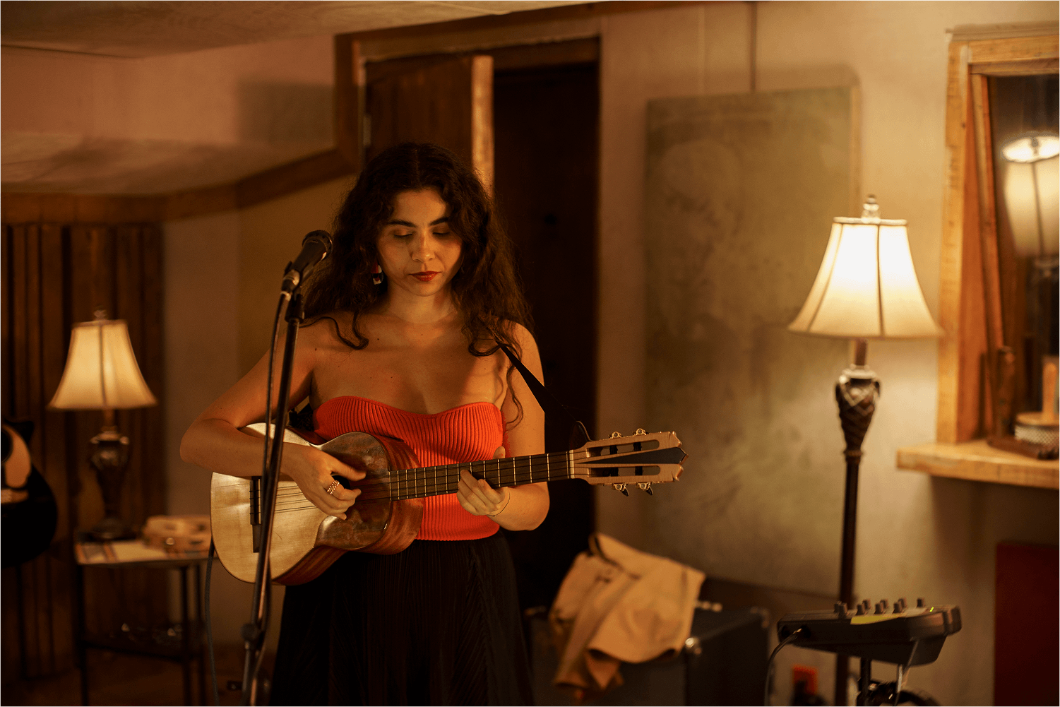 Woman in a red top playing an acoustic guitar indoors with warm lighting and a microphone in front.