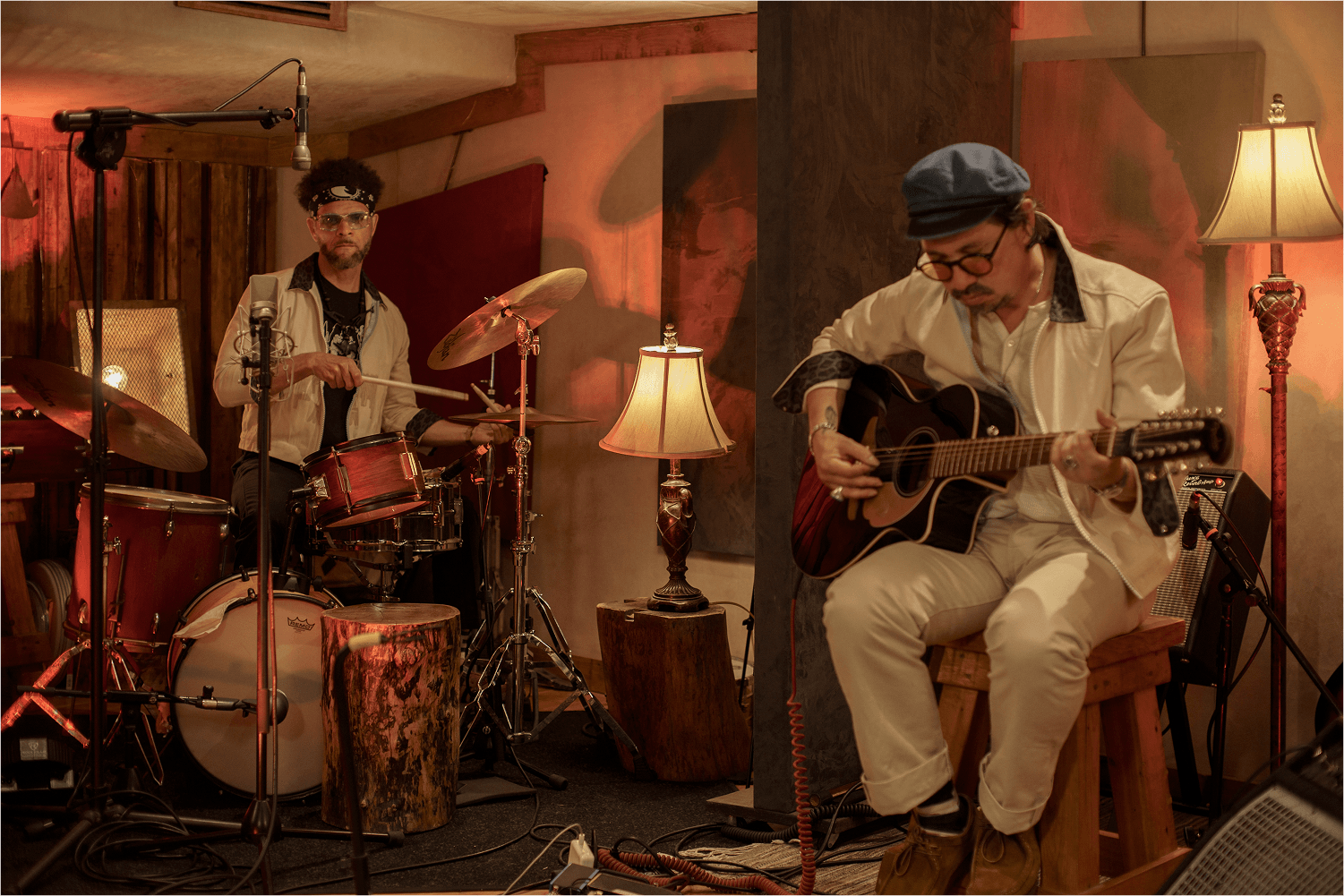 Two musicians in a warmly lit studio, one playing drums and the other playing an acoustic guitar seated on a wooden stool.
