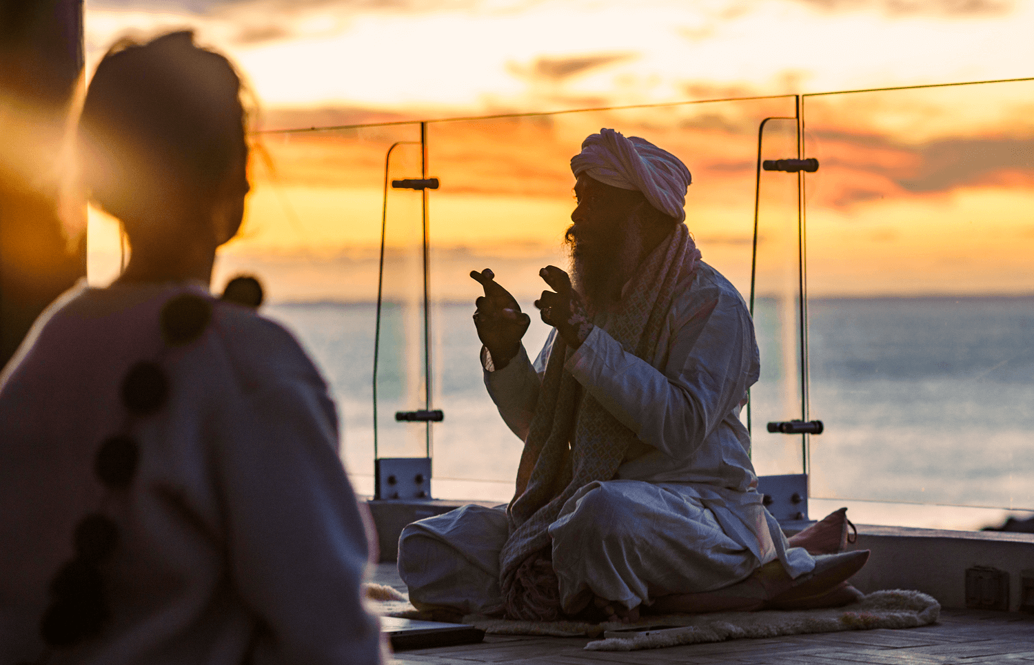 Silueta de un hombre con traje tradicional meditando al atardecer cerca de una barandilla de cristal junto al mar, con otra persona borrosa en primer plano.