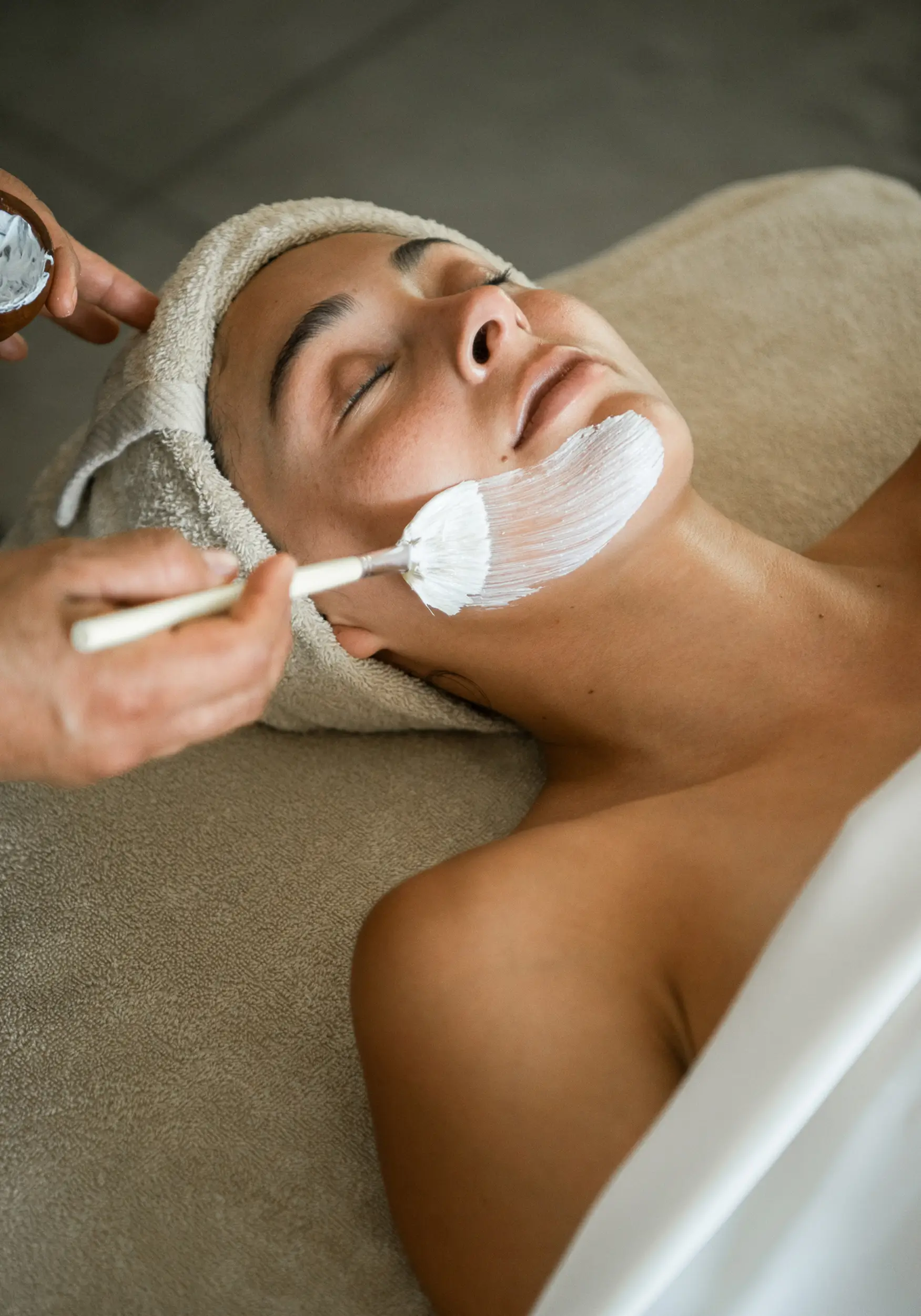 Woman with a towel wrapped around her head receiving a white facial mask applied to her chin while lying on a spa bed.