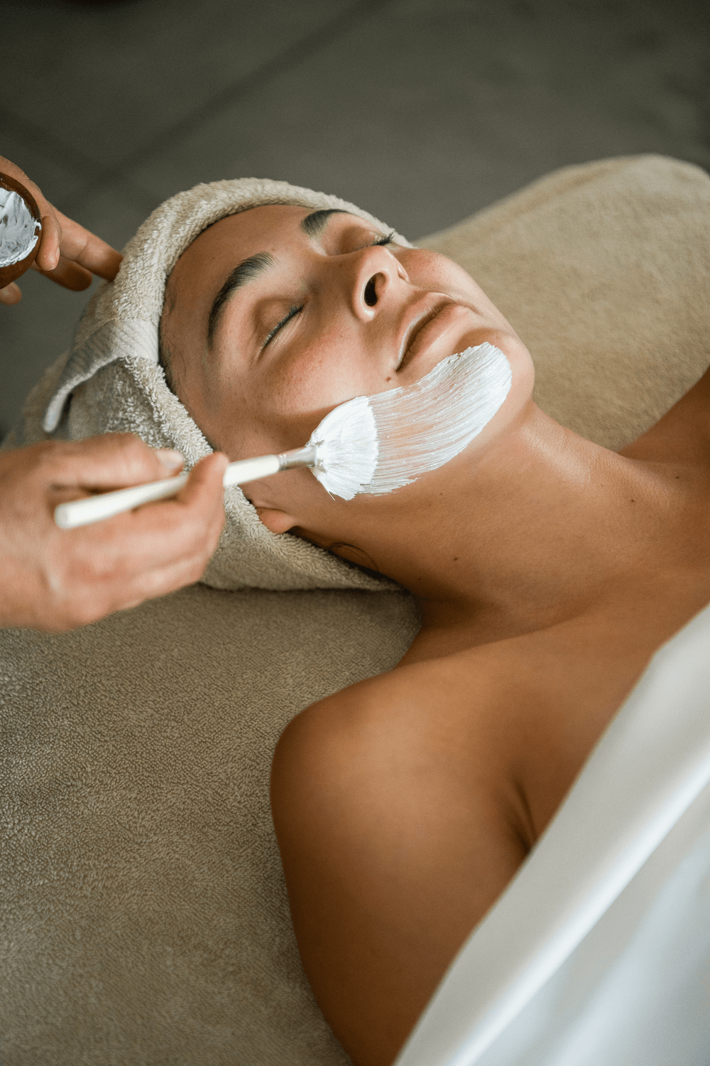 Mujer con una toalla envuelta alrededor de la cabeza recibiendo una mascarilla facial blanca aplicada en la barbilla mientras está tumbada en una camilla de spa.