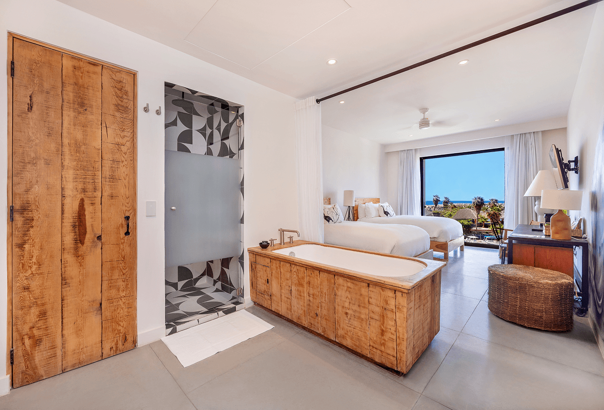 Habitación de hotel moderna con dos camas, bañera de madera, ducha con motivos decorativos y gran ventana con vistas al mar.