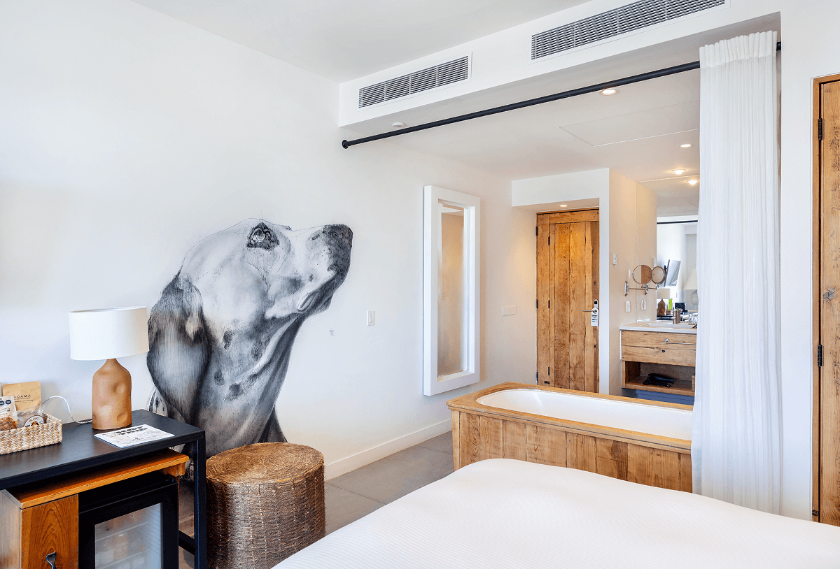 Dormitorio moderno con una cama blanca, una bañera de madera, puertas de madera natural y un mural en la pared con la cabeza de un perro mirando hacia arriba.