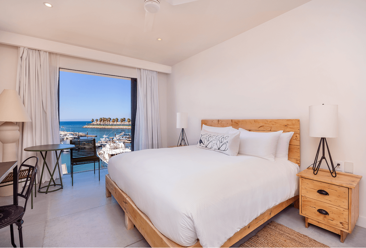 Modern hotel room with a king-size bed, wooden frame, two bedside tables with lamps, and a balcony window showing a marina and ocean view.