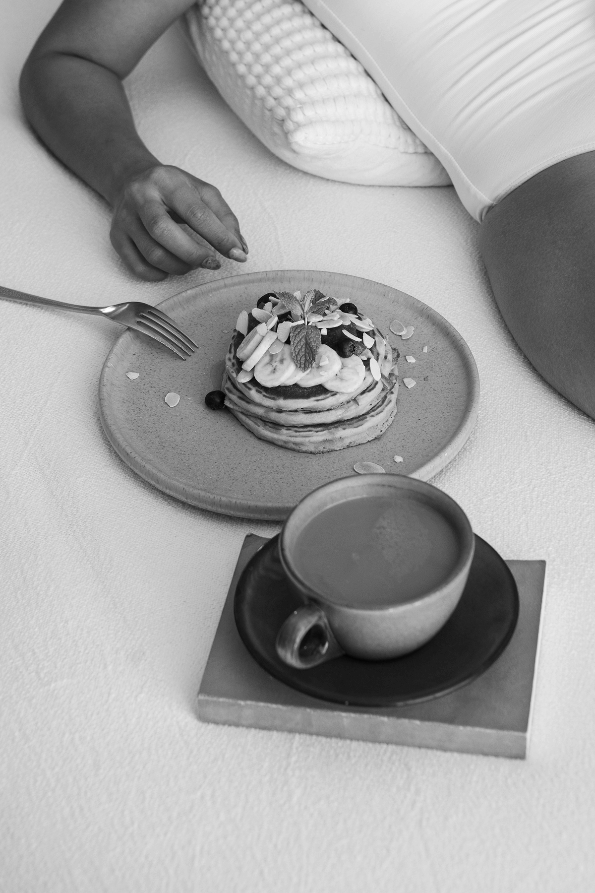 Persona tumbada en una cama con un plato de tortitas cubiertas con rodajas de plátano, arándanos, almendras laminadas y hojas de menta, junto a una taza de café en un platillo.