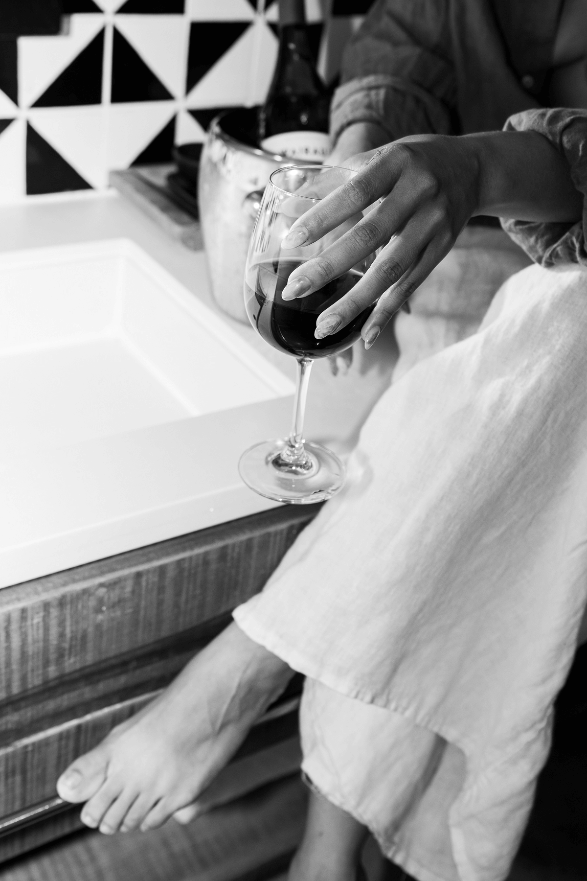 Persona sentada descalza sobre un armario de madera sosteniendo una copa de vino tinto cerca de un fregadero con azulejos geométricos en la pared al fondo.