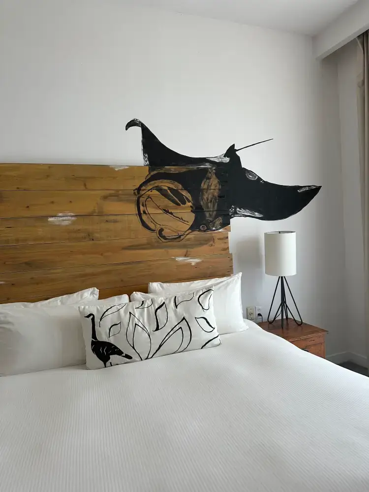 Bed with white linens, wooden headboard featuring a black dog painting, and a black and white pillow with leaf and bird designs next to a wooden nightstand with a lamp.