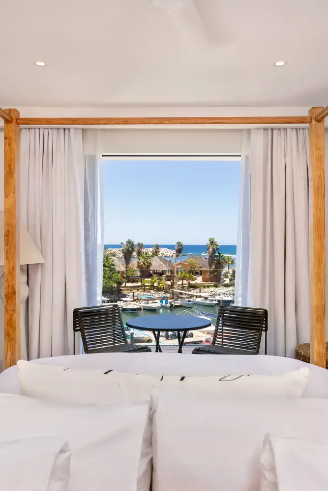 View from a bedroom with white bed and curtains looking out onto a balcony with two chairs and a table, overlooking boats, palm trees, and the ocean.